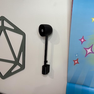 May include: A black, cylindrical object with a long, thin stem and a rectangular base, mounted on a white wall. The object has a circular opening on one end. A geometric, gray-green shape is on the left.