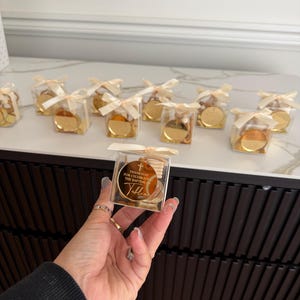 May include: A white ceramic mug with a black handle and the text "But First, Coffee" in a bold, black font. The mug is set against a blurred background, suggesting a cosy setting.