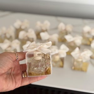 May include: Small, clear glass bottles filled with a golden liquid, each presented in a clear box and tied with a cream-coloured bow. The bottles have a gold label with the words "WELCOME BABY" and a name. A hand holds one of the bottles.
