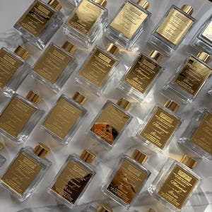 May include: A collection of perfume bottles with gold detailing. The clear, rectangular bottles have gold caps and labels with the text "Mohamed Feras". They are arranged on a white marble surface, creating a luxurious aesthetic.