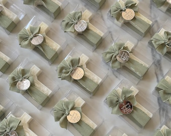 May include: A collection of cross-shaped items packaged in clear plastic boxes, each adorned with a sage green fabric ribbon and a circular metallic tag engraved with the text Baptism of Gabriel and a date.