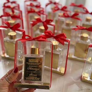May include: Clear glass bottles with gold lids and labels, presented in clear boxes tied with red satin bows. Each bottle has Arabic script and English text on the gold label. The bottles are arranged on a white surface.