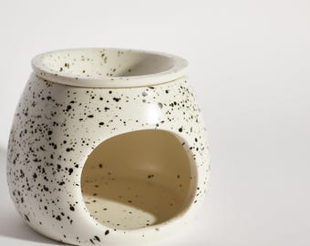 May include: White ceramic wax melt burner with black speckled design. The burner has a round, open top and a large, open well in the side.