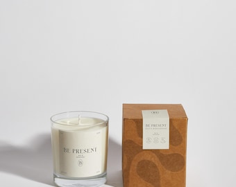 May include: A brown cardboard box with a white label that reads "BE PRESENT" and a small logo. The box is sitting next to a clear glass candle jar with a white candle inside. The candle jar also has a white label that reads "BE PRESENT".