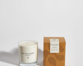 May include: A brown cardboard box with a white label that reads "ON VACATION CALM OF SOLITUDE" and a white circle with the letter "B" inside. The box is sitting on a white surface next to a clear glass candle jar with a white candle inside. The candle jar has a white label that reads "ON VACATION CALM OF SOLITUDE" and a white circle with the letter "B" inside.