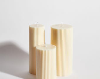 May include: Three off-white pillar candles with a ribbed texture. The candles are arranged in a row, with the largest candle in the back and the smallest candle in the front.
