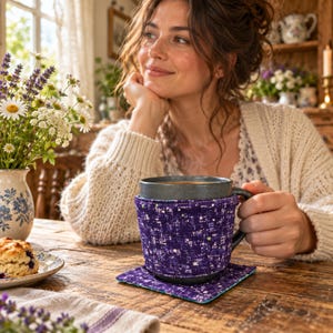 Puede incluir: Una taza de cerámica gris oscuro con una funda de tela morada con un patrón de motas blancas y verde claro, apoyada sobre un posavasos de tela cuadrado a juego. La funda envuelve la taza, proporcionando una apariencia texturizada.