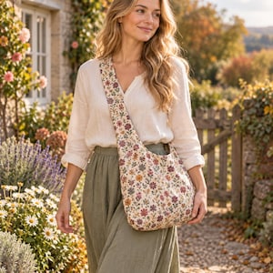 Könnte beinhalten: Eine Umhängetasche mit cremefarbener Basis und einem dichten, mehrfarbigen Blumenmuster in Rot-, Orange-, Lila- und Grüntönen. Die Tasche verfügt über einen breiten, integrierten Schulterriemen und eine weiche, unstrukturierte Form, die auf der Hüfte liegt.