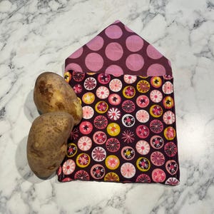 Könnte beinhalten: Eine wiederverwendbare Kartoffelaufbewahrungstasche aus Stoff mit Klappe, mit einem dunkelvioletten Blumenmuster und einem gepunkteten Oberteil. Zwei ungeschälte Kartoffeln liegen neben der Tasche auf einer Marmoroberfläche. Die Tasche ist für den Küchengebrauch konzipiert.