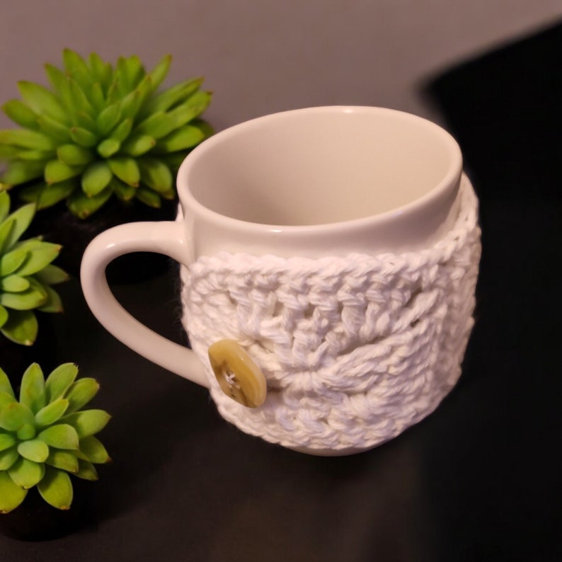May include: White ceramic mug with a white crocheted cosy featuring a button. The cosy is a simple stitch pattern.