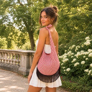 May include: A red and white gingham shoulder bag with a black fringe trim. The bag is worn over the shoulder and has a curved bottom. The background features a stone wall, lush greenery, and a person.