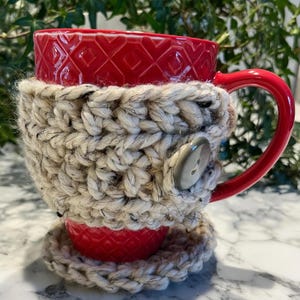 May include: A red ceramic mug with a textured diamond pattern, wrapped in a beige and black speckled crocheted cozy. The cozy has a large gray button closure and sits on a matching coaster. The mug has a red handle.