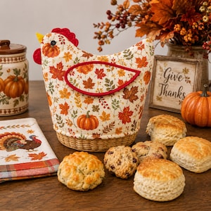 May include: A fabric chicken-shaped tea cosy featuring a pattern of orange pumpkins and autumn leaves in shades of red, yellow, and brown. The cosy has a red comb, a yellow beak, and a woven base, surrounded by several baked biscuits on a table.