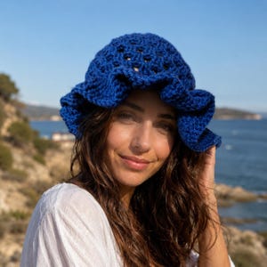 May include: A blue crochet hat with a ruffled brim. The hat is made of a textured yarn and has a slightly open weave. The hat is worn by a person with long, dark hair and a white top. The background features a body of water and a blue sky.