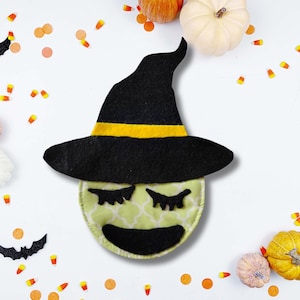 May include: A Halloween-themed felt witch face with a black hat and yellow band. The witch face has closed eyes and a smiling mouth. The background includes pumpkins, confetti, and candy corn.