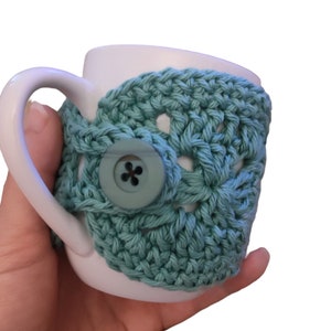 May include: A white ceramic mug with a teal crocheted cosy. The cosy has a button closure and a crocheted design.