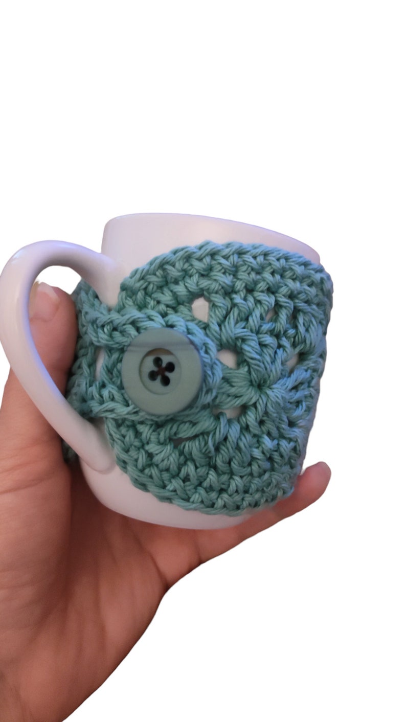 May include: A white ceramic mug with a teal crocheted cosy. The cosy has a button closure and a crocheted design.