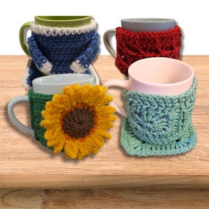 May include: Four mugs with colourful, crocheted cosies. One mug has a blue and white cosy, another a red cosy, a third has a green cosy with a sunflower, and the last has a blue and green cosy. The mugs are on a wooden surface.