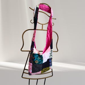 May include: A patchwork shoulder bag with a pink lining and strap. The bag features a variety of patterned fabrics in pink, black, white, and green. The bag is displayed on a gold-coloured metal stand.
