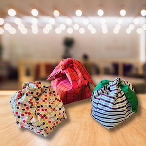 May include: Three fabric gift bags with different patterns. The bags are tied with a knot at the top and are made of a soft, colorful fabric. The bags are on a wooden surface.