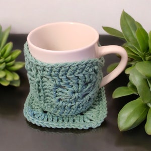 May include: A white ceramic mug with a teal crocheted mug cozy. The cozy has a square shape with a crocheted design.