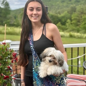 May include: A woman with long brown hair is holding a small white dog in a colorful patterned sling bag. The dog is looking at the camera. The woman is wearing a black tank top and black pants. The background is a blurry green field and a white fence.