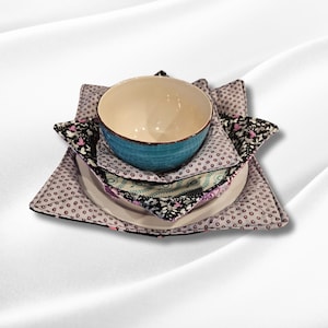 Puede incluir: Un conjunto de vajilla que incluye un cuenco azul claro, un plato blanco y un salvamanteles de tela estampada. El cuenco tiene un exterior texturizado y un interior color crema. El salvamanteles presenta un diseño floral y de lunares.