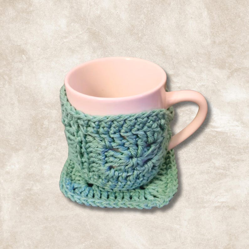 May include: A crocheted mug cosy in shades of teal and blue, with a pink ceramic mug inside. The cosy is made with a square granny stitch pattern.