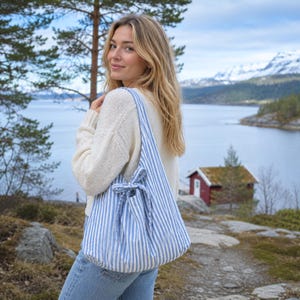 Op de afbeelding: Een blauw-wit gestreepte draagtas met een trekkoordsluiting wordt over de schouder gedragen. De tas is gemaakt van een zachte stof en heeft een casual, relaxte stijl. De achtergrond toont een schilderachtig uitzicht op een meer en bergen.