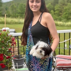 Puede incluir: Una bolsa de transporte para mascotas estampada en tonos azules, morados y amarillos, que sostiene un perro pequeño blanco y negro. La bolsa se lleva al hombro. El fondo incluye una persona, una terraza y vegetación.