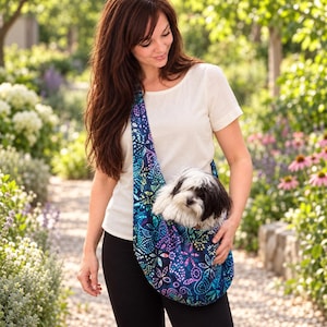 Könnte beinhalten: Eine Haustiertragetasche aus Stoff mit einem lebendigen, mehrfarbigen Blumen- und Paisley-Muster in Blau-, Lila- und Rosatönen. Die Tasche wird quer über dem Körper getragen und enthält einen kleinen, flauschigen schwarz-weißen Hund.