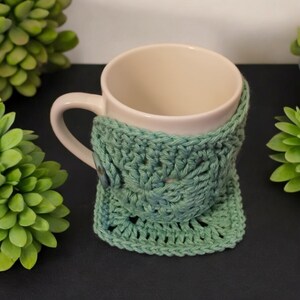 May include: A white ceramic mug with a teal crocheted cosy. The cosy has a button on the side.