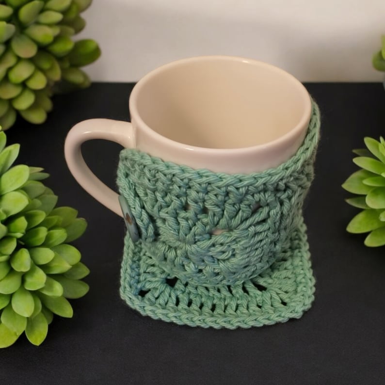 May include: A white ceramic mug with a teal crocheted cosy. The cosy has a button on the side.