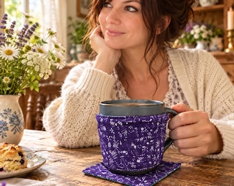 Puede incluir: Una taza de cerámica gris oscuro envuelta en una funda de tela acolchada de color morado con un patrón floral blanco, apoyada sobre un posavasos de tela cuadrado a juego. La taza tiene un asa resistente y está sobre una superficie de madera.