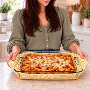 Peut inclure: Un plat de cuisson rectangulaire en verre rempli de pâtes penne à la sauce tomate gratinées au fromage, placé dans un support en tissu jaune et blanc à motifs floraux doté de poignées latérales pour faciliter le transport.