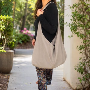 May include: A large, beige shoulder bag made of soft fabric. The bag is worn over the shoulder and hangs down. The bag is a neutral color and appears to be made of linen or a similar material. The bag is a simple, casual style.