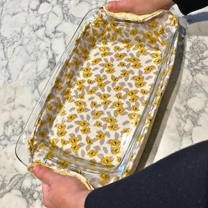 May include: A clear glass rectangular baking dish with a floral patterned fabric cover. The fabric features yellow flowers and gray leaves on a cream background. The dish is being held by two hands.