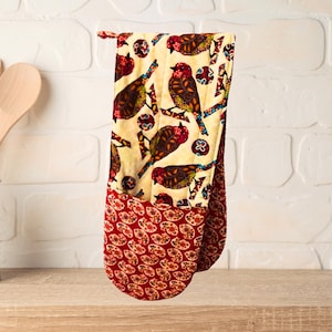 May include: A double oven mitt with a bird pattern. The top portion has a cream background with brown and red birds. The bottom portion is red with a floral pattern. The mitt is hanging on a wooden surface.