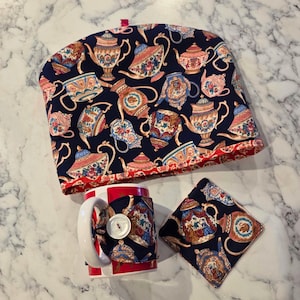 May include: A navy blue tea cozy, mug wrap, and coaster set. The set features a repeating pattern of ornate teapots in shades of pink, blue, and gold. The mug is red and white striped with a white handle. A white button secures the mug wrap.