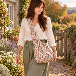 May include: A crossbody bag featuring a cream-colored fabric with a dense, repeating floral pattern in shades of muted red, dusty pink, and sage green. The bag has a wide, integrated shoulder strap and a soft, unstructured, slouchy silhouette.