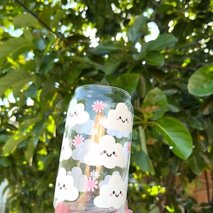 May include: A clear glass can with a white and pink cloud pattern. Each cloud has a smiling face and pink flowers.