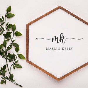 May include: A white, hexagon-shaped sign with a brown wooden frame. The sign features the initials "mk" in elegant script above the name "MARLIN KELLY" in a simple font. Green leafy branches are on the left side of the sign.