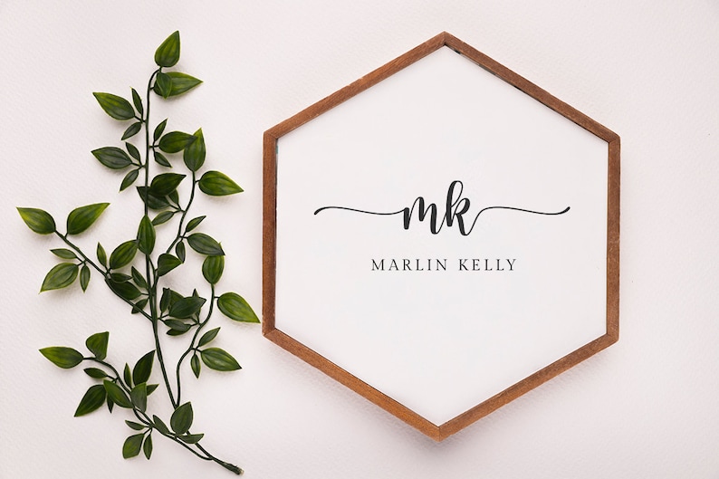 May include: A white, hexagon-shaped sign with a brown wooden frame. The sign features the initials "mk" in elegant script above the name "MARLIN KELLY" in a simple font. Green leafy branches are on the left side of the sign.