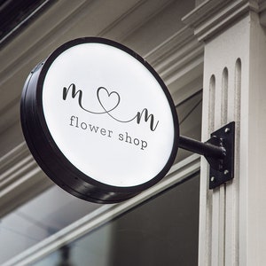 May include: A round, illuminated flower shop sign with a black frame. The sign displays the text "m♡m flower shop" in a delicate, handwritten font. The sign is mounted on a black metal bracket attached to a white building.