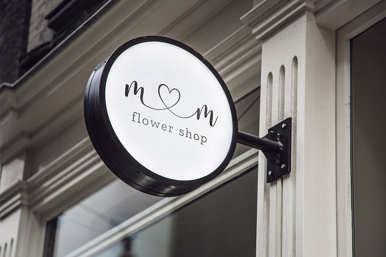 May include: A round, illuminated flower shop sign with a black frame. The sign displays the text "m♡m flower shop" in a delicate, handwritten font. The sign is mounted on a black metal bracket attached to a white building.