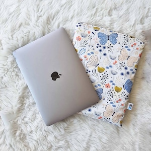 May include: A silver laptop with a black Apple logo rests next to a white laptop sleeve. The sleeve features a floral and butterfly pattern in blue, pink, and yellow. The sleeve is designed to protect the laptop.