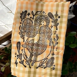 May include: A fabric square with a fox illustration in brown and black, surrounded by leaves and botanical elements. The fabric has a yellow and orange gingham pattern. The text "H.E. creations" is visible.