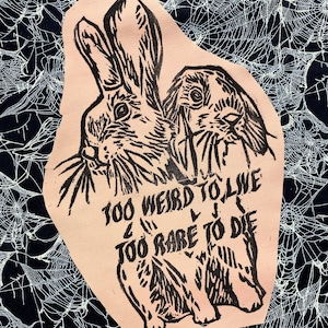 May include: A light-colored patch featuring a black ink block print of two rabbits. The text "TOO WEIRD TO LIVE, TOO RARE TO DIE" is printed in black ink below the rabbits. The design is set against a black background with a white spiderweb pattern.
