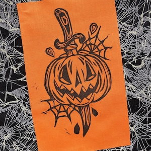 May include: A black and white linocut print of a pumpkin with a knife through it. The pumpkin has a carved face and is surrounded by spiderwebs.