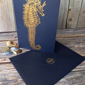 May include: A navy blue card with a gold foil embossed seahorse design. The card is blank inside and comes with a matching navy blue envelope.